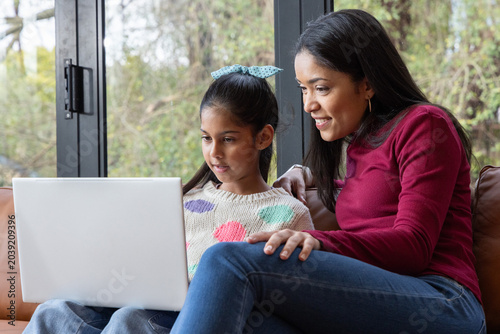 Indian mother leaning over school-aged daughter, helping with white laptop on leather sofa at home