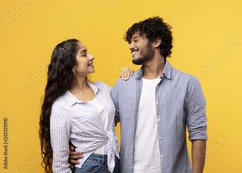 Obraz na płótnie Joyful South Asian Couple Smiling While Looking At Each Other, Standing Together Against A Bright Yellow Background
