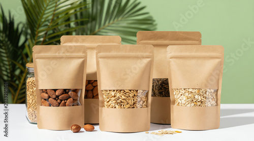 Various nuts seeds and oats in eco friendly kraft paper pouches on white table with green plant. Healthy snacks in compostable packaging ready for sale or consumption.