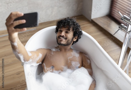 Tableau sur toile Happy Young Man Of South Asian Descent Smiling While Taking Selfie In A Bathtub Filled With Bubbles, Relaxing And Enjoying Spa-like Atmosphere At Home, Self-care And Wellness Concept