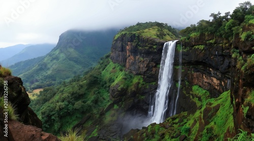 Water cascades down steep green mountain cliff in India. Lush vegetation covers rocky hillsides. Mist rises from plunging water in a tropical valley. Serene natural landscape.