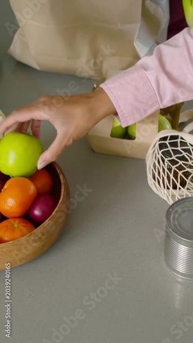 Vertical video: Hand reaching net bag, person in pink moving apples from tray in kitchen, unpacking