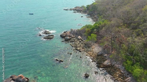 Aerial view of a dense, vibrant green coastal jungle meeting dark blue ocean water. A thin, rocky beach and white foamy waves separate the rugged terrain from the sea.concept	