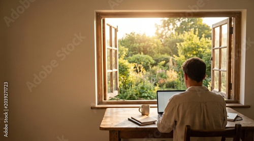 Man working on laptop at wooden desk by open window with sunlit garden view