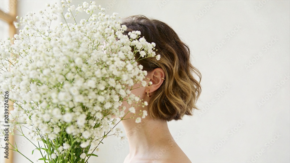Naklejka premium Young woman with short wavy hair holding large bouquet of white baby's breath flowers covering face, minimalist style, soft lighting, natural beauty, floral arrangement, spring