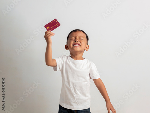 Happy young asian boy holds a red credit card near his ear while smiling broadly during this bright studio photo that captures a playful moment of childhood financial learning experiences.