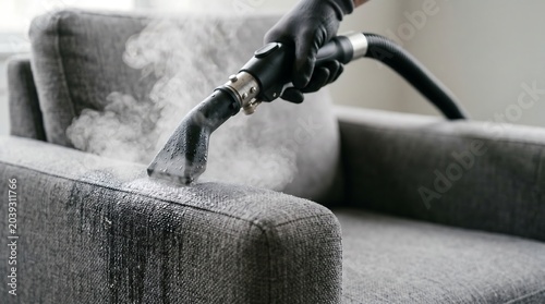 Professional cleaner using a hot steam cleaning machine on a grey fabric armchair for deep disinfection and stain removal