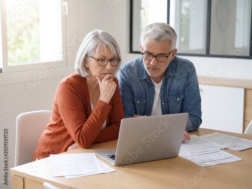 A serious middle aged couple sitting at a wooden table at home looking at their laptop screen while reviewing various financial documents and bills