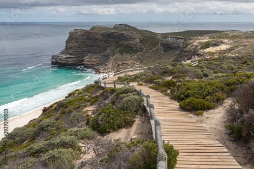 Konstfotografi Scenic view of Cape of Good Hope, South Africa, featuring a wooden boardwalk trail with handrails leading towards a dramatic rocky cliff, turquoise ocean water, and a cloudy sky over Dias Beach