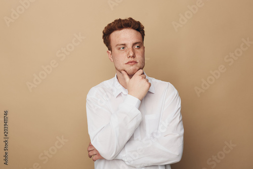 фотографія Thoughtful young man in white shirt looks away with hand on chin against beige background