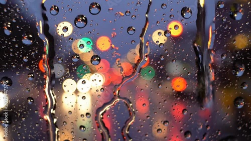 Raindrops cling to a clear glass surface while vibrant bokeh lights glow in the dark evening background.