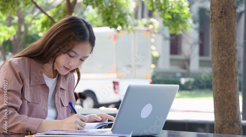 Foto Young Thai University Student Writing in Notebook with Laptop Outdoors