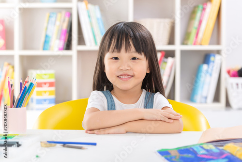 Quadro em tela A polite and happy little Asian girl sitting up straight at her desk with crossed arms and smiling brightly at the camera
