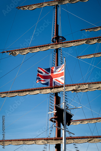Great britain union jack flag waving on a mast with a replica sailboat mast in the background.