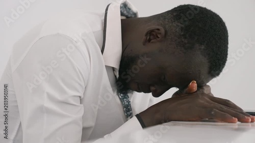 Defeated man with head bowed. Black man sits postdate in private remorse, hands folded on table, heavy breathing and introspective silence white shirt and tie, intimate closeup of exhaustion, grief