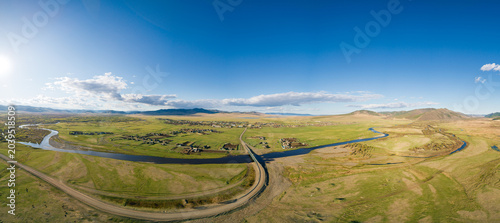 Aerial panoramic of a winding river in green plains near a village, road, and bridge under a brilliant blue sky