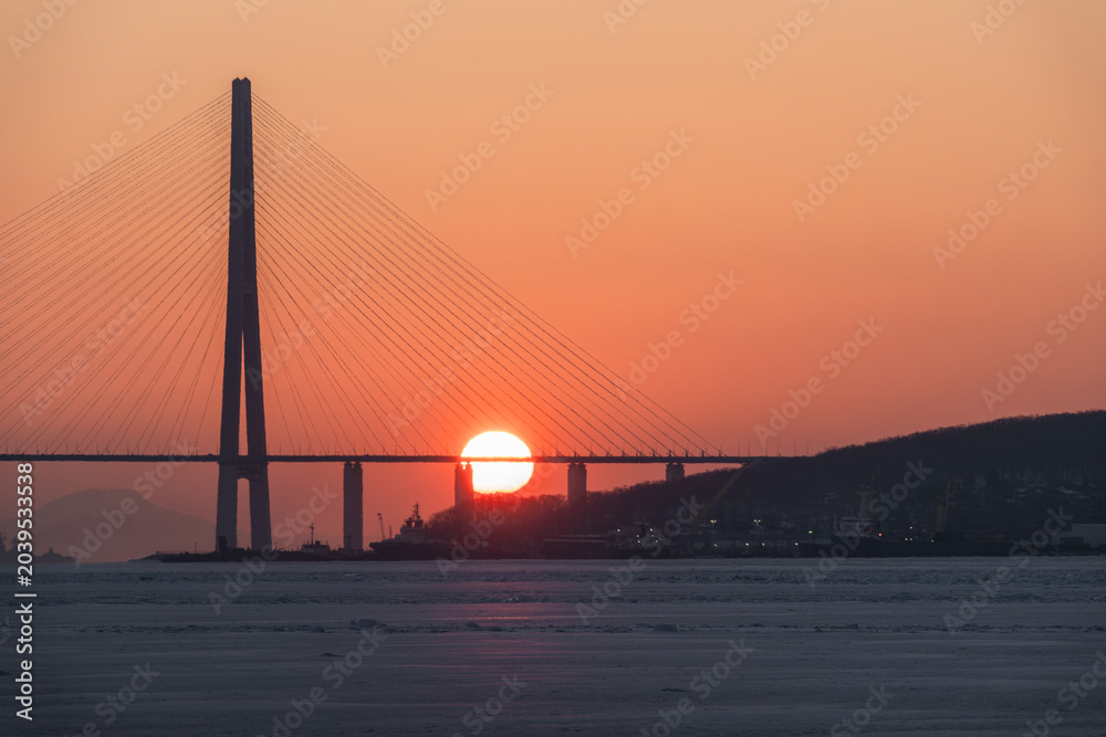 Obraz premium Sunset view behind a suspension bridge with cables silhouetted against an orange sky over a calm body of water reflecting the warm hues of dusk