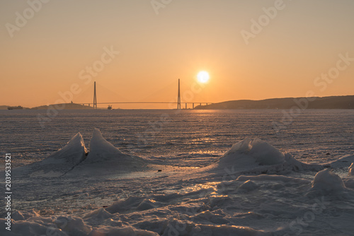 Bild auf Leinwand Sunset over frozen river with ice formations and bridge silhouette in the backgr
