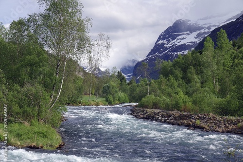 A clear river rushes through a mountain and forest landscape with snow-capped peaks in the background, Loen, Nordfjord, Norway