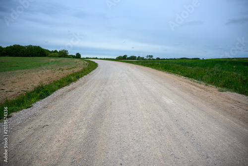 Country dirt road stretching through rural landscapes and green meadows on a cloudy day.