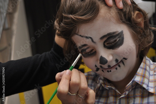 Child with skull makeup being painted during Halloween preparation at a local event in afternoon light