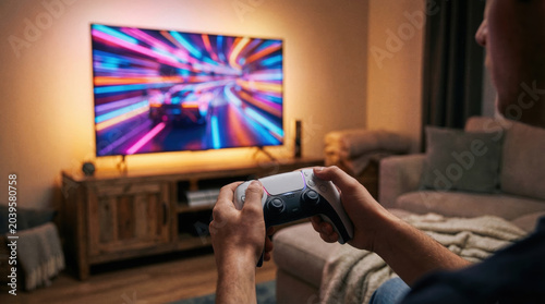 Over The Shoulder View Of Man Holding Wireless Controller Playing Video Game In Ambient Lit Living Room