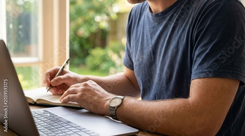 Professional man working on silver laptop while taking notes in notebook at wooden desk with blurred background inside home office setting during bright daylight