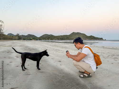 Man crouching to photograph a curious black dog on a peaceful sandy beach at low tide, with distant mountains