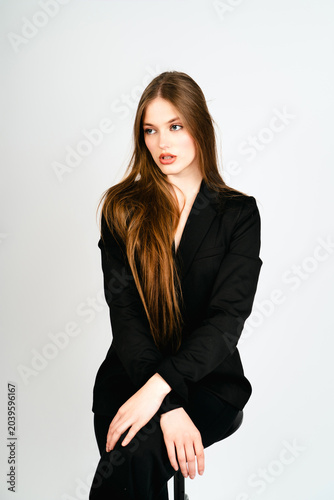 Foto Pensive female model in a black pantsuit sitting on a high stool and looking away in a studio with a white backdrop