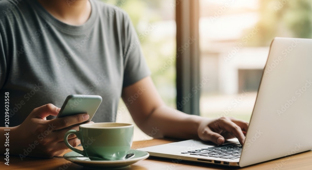 Fototapeta premium Woman sitting near window with coffee cup and smartphone while typing on laptop computer working remotely in cozy cafe environment with blurred natural background