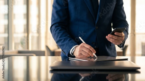 Business executive in formal suit writing on digital tablet with stylus pen and holding smartphone while working at modern office desk with reflection on glass top