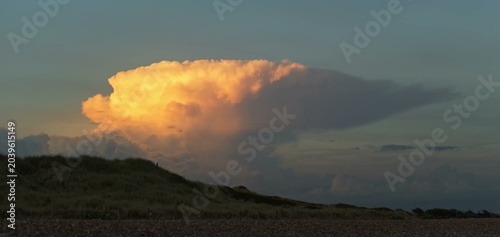 Картината върху платно A massive cumulus cloud glows with intense orange light against a darkening twilight sky as it towers over a silhouette of grassy coastal dunes and a pebbled beach shoreline