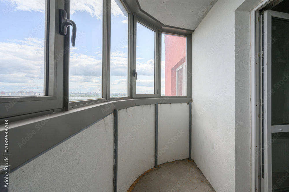 Obraz premium curved balcony with grey-framed panoramic windows, white insulated walls, and a bare concrete floor, offering a view of the sky and neighboring buildings