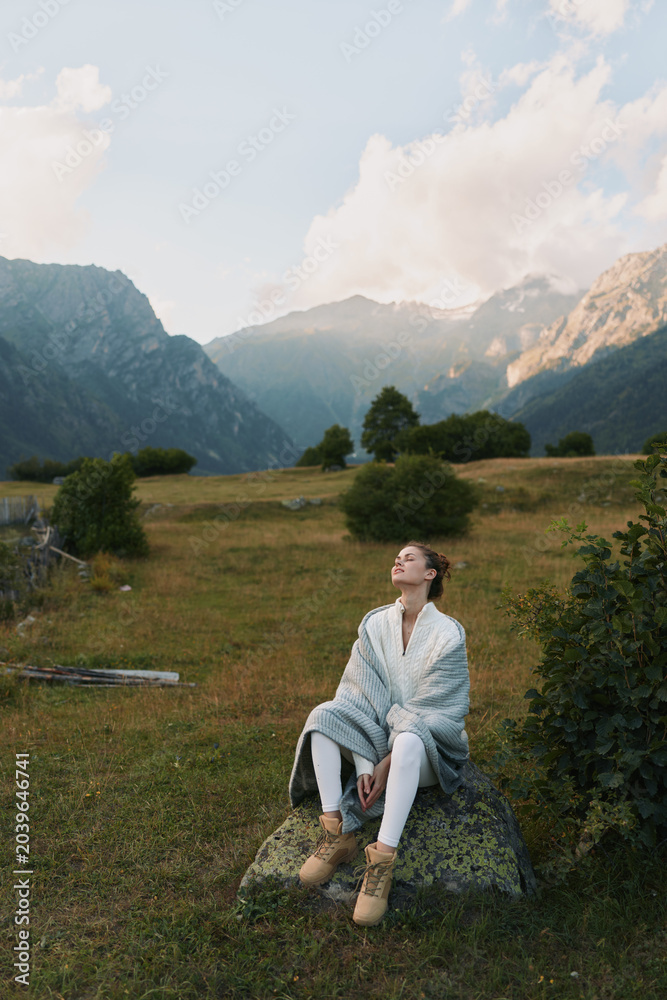 Fototapeta premium Young woman sits on a rock in a grassy meadow with distant mountains, savoring a peaceful outdoor moment, scene filled with soft light, warm tones and quiet contemplation