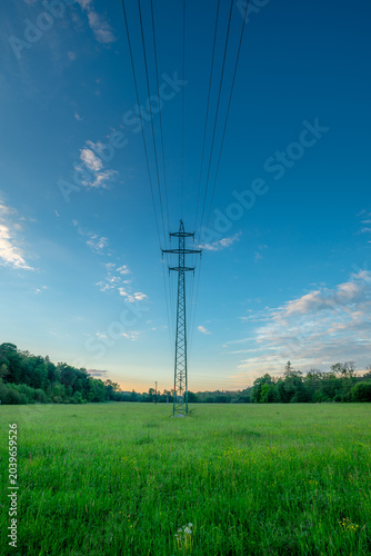 Tableau sur toile Tall steel electrical power transmission tower situated in a lush green countryside field, with thick overhead cables stretching across a bright blue sky during a peaceful summer day
