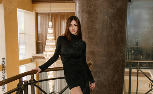 A beautiful girl model in a black tight dress on a wrought-iron balcony with a huge chandelier behind her.