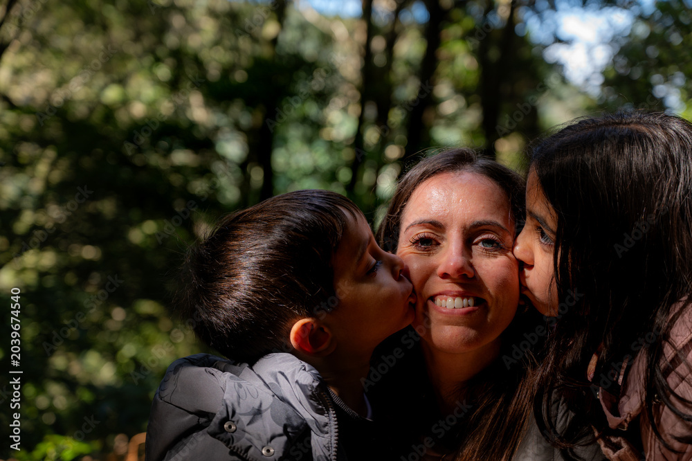Fototapeta premium The mother smiles tenderly as her children kiss her on the cheeks. Sunlight filters through the trees behind them.