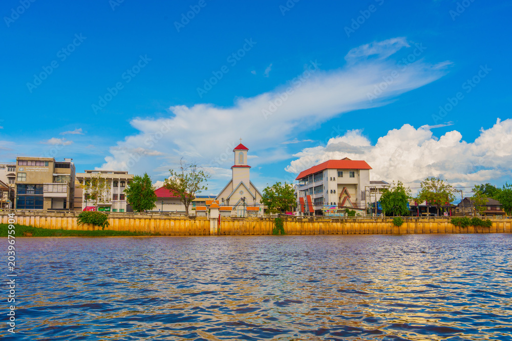 Obraz premium Riverside town with church tower, residential buildings, and blue sky with clouds reflecting on water surface in Chiang Mai Thailand