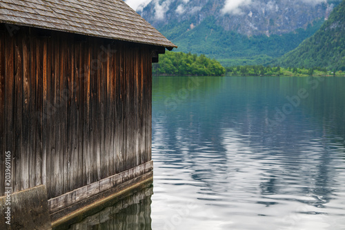 Картината върху платно Rustic lakeside boathouse with calm water reflections and alpine mountains under cloudy sky, Hallstatt, Austria