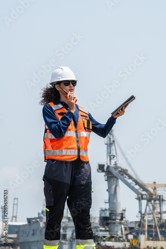 Woman wears a white hard hat and high-visibility safety vest. She holds a tablet and speaks on a mobile phone. Industrial cranes and shipping vessels fill the background