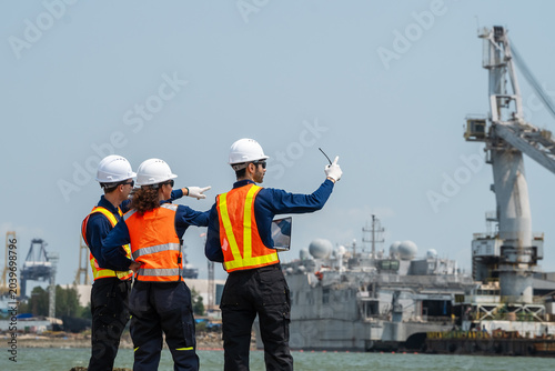 Three logistics engineers study port infrastructure. They wear white hard hats and safety vests. One points toward the harbor area. Cranes and ships dominate the background