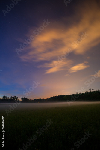Fotografija A thick fog settling over a vast, quiet meadow under a dramatic, glowing sunset sky that is slowly fading into the darkness of night