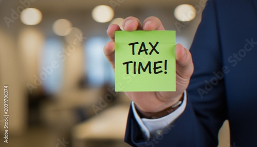 A business professional in a suit holding a green sticky note with the words Tax Time written on it in an office setting.