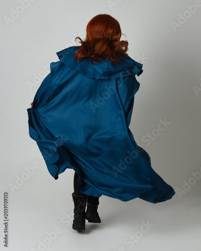 full length portrait of female model wearing medieval fantasy warrior historical costume with blue hooded cloak. silhouette figure, walking away back view on studio background.