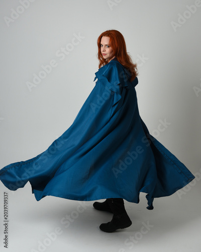full length portrait of female model wearing medieval fantasy warrior historical costume with blue hooded cloak. silhouette figure, walking away back view on studio background.