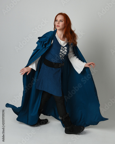 full length portrait redhead female model wearing medieval fantasy warrior historical costume with blue hood cloak. walking away with magical spell arm gesture, silhouette figure on studio background.