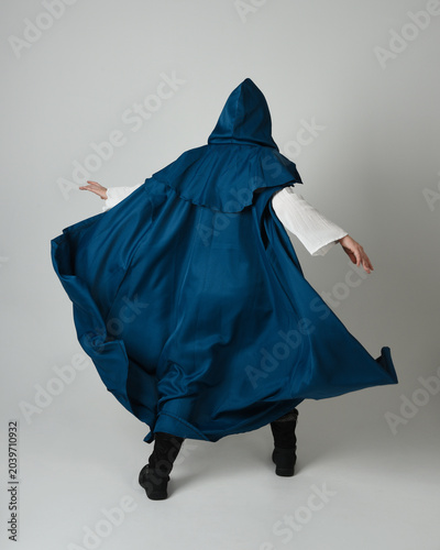 full length portrait of model wearing medieval fantasy warrior historical costume with blue hooded cloak. silhouette figure,  walking away in back view on studio background.