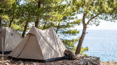 Two beige camping tents set up near a rocky shoreline surrounded by pine trees with a clear view of the ocean and bright blue sky in the background
