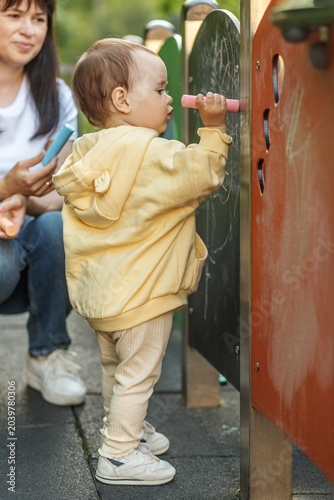 Toddler drawing with chalk while mother supervises outdoor playground activity