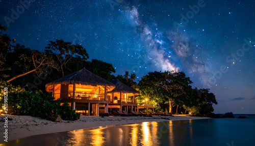 Luxury tropical eco resort bungalows on beach under beautiful starry night sky with Milky Way galaxy reflection on water